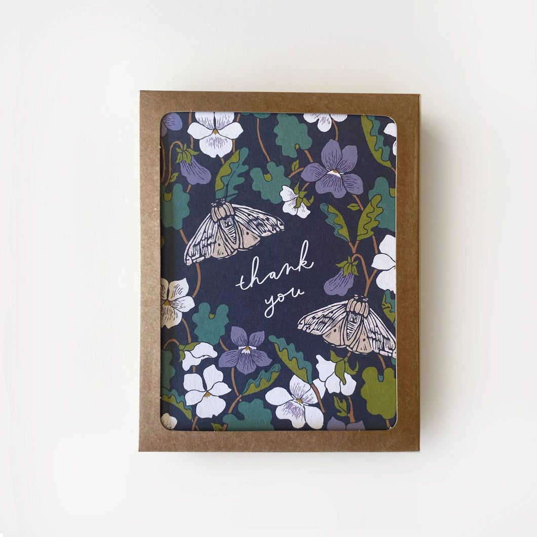 Woodland greeting card with a slate background color, with intermingling purple and white flowers, green leaves, and two beautiful moths.  The card reads thank you in cursive white writing in the center.